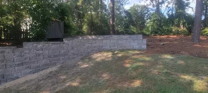 A retaining wall in a yard, with steps on the left and mulch and trees in the background.