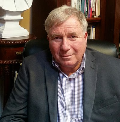 A man in a suit and plaid shirt is sitting in front of a bookshelf