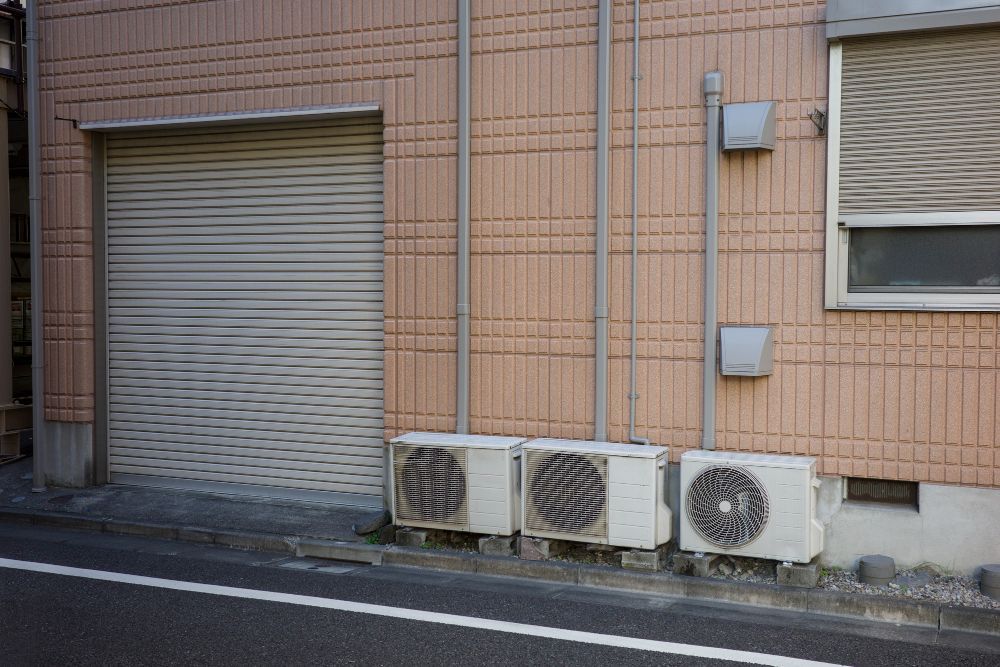 A building with a garage door and air conditioners on the side of it.