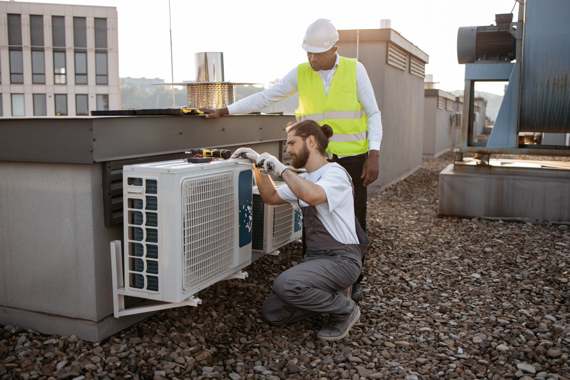 Two men are working on an air conditioner on the roof of a building.