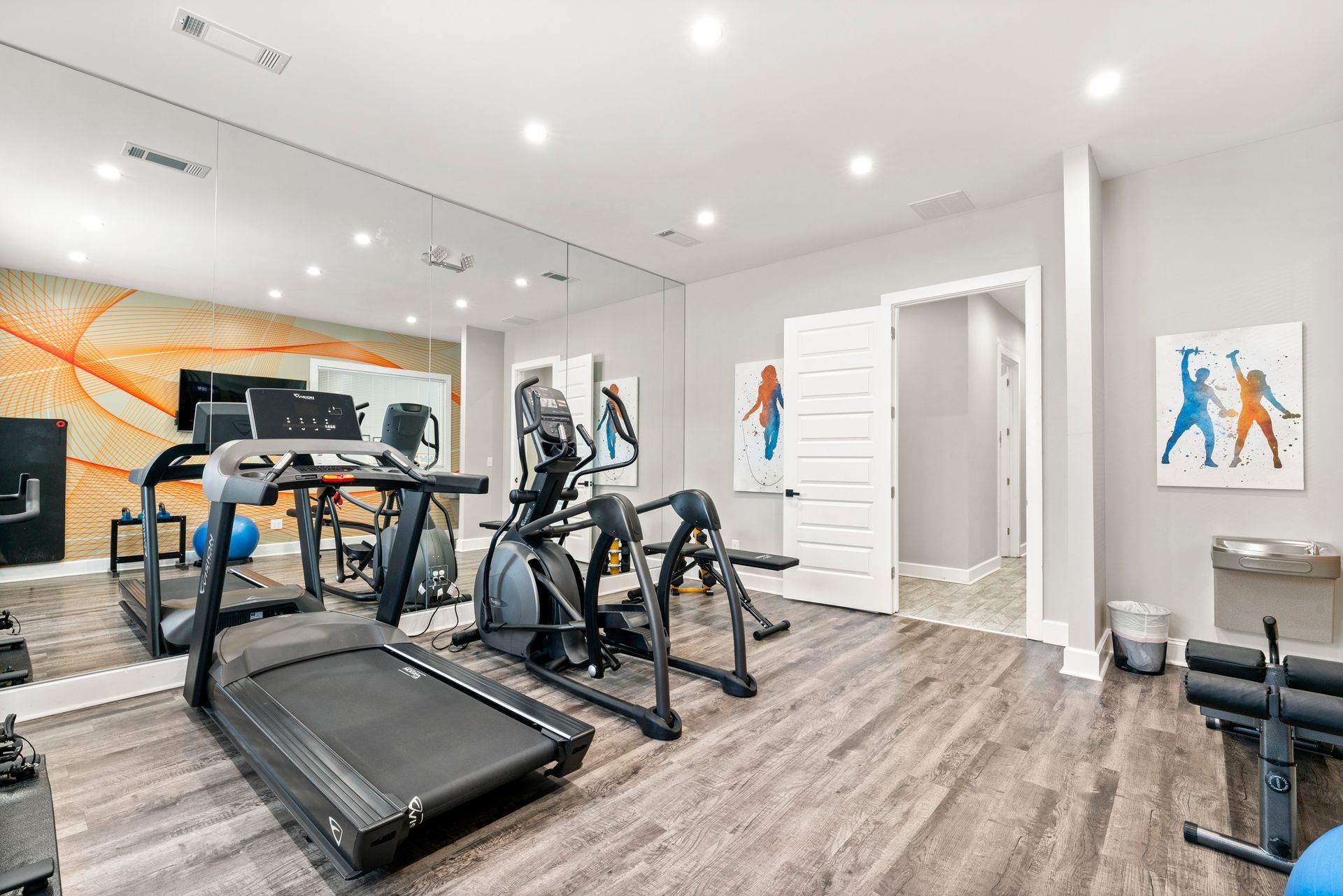 Modern home gym with treadmill, elliptical, exercise bike, mirrored wall, and light wood flooring