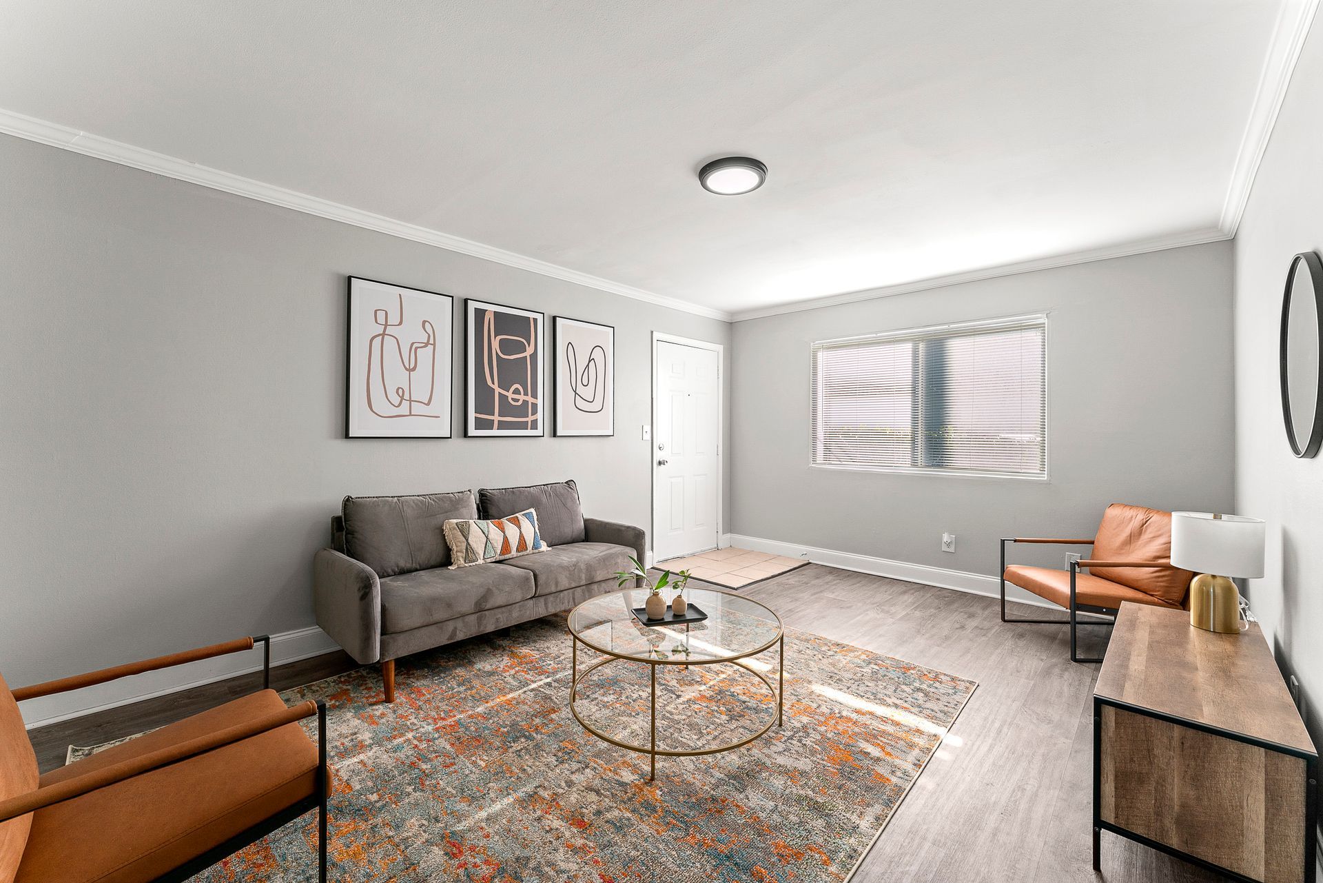 Bright living room with gray sofa, patterned rug, round coffee table, TV console, and window blinds