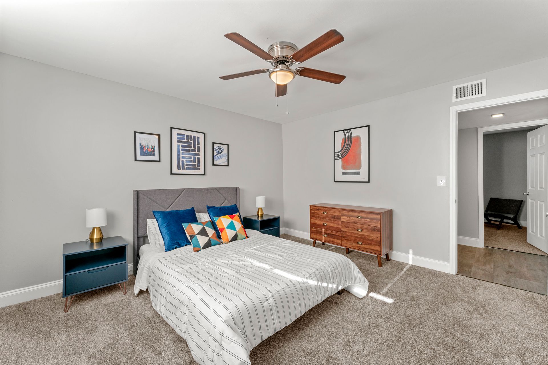Bedroom with a gray bed, blue nightstands, wall art, ceiling fan, and beige carpet.
