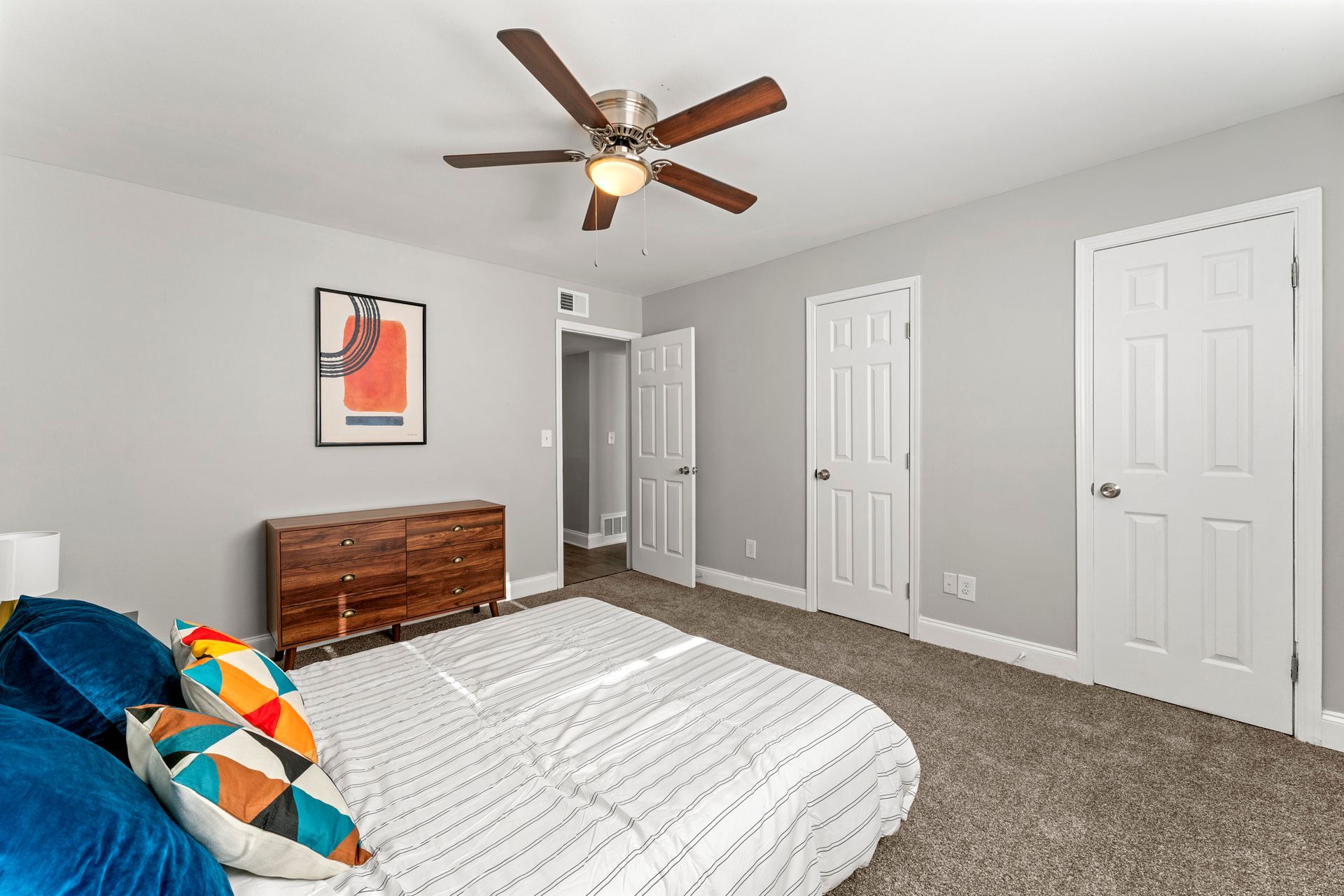 Bright bedroom with white walls, ceiling fan, bed, wood dresser, and two white closet doors.