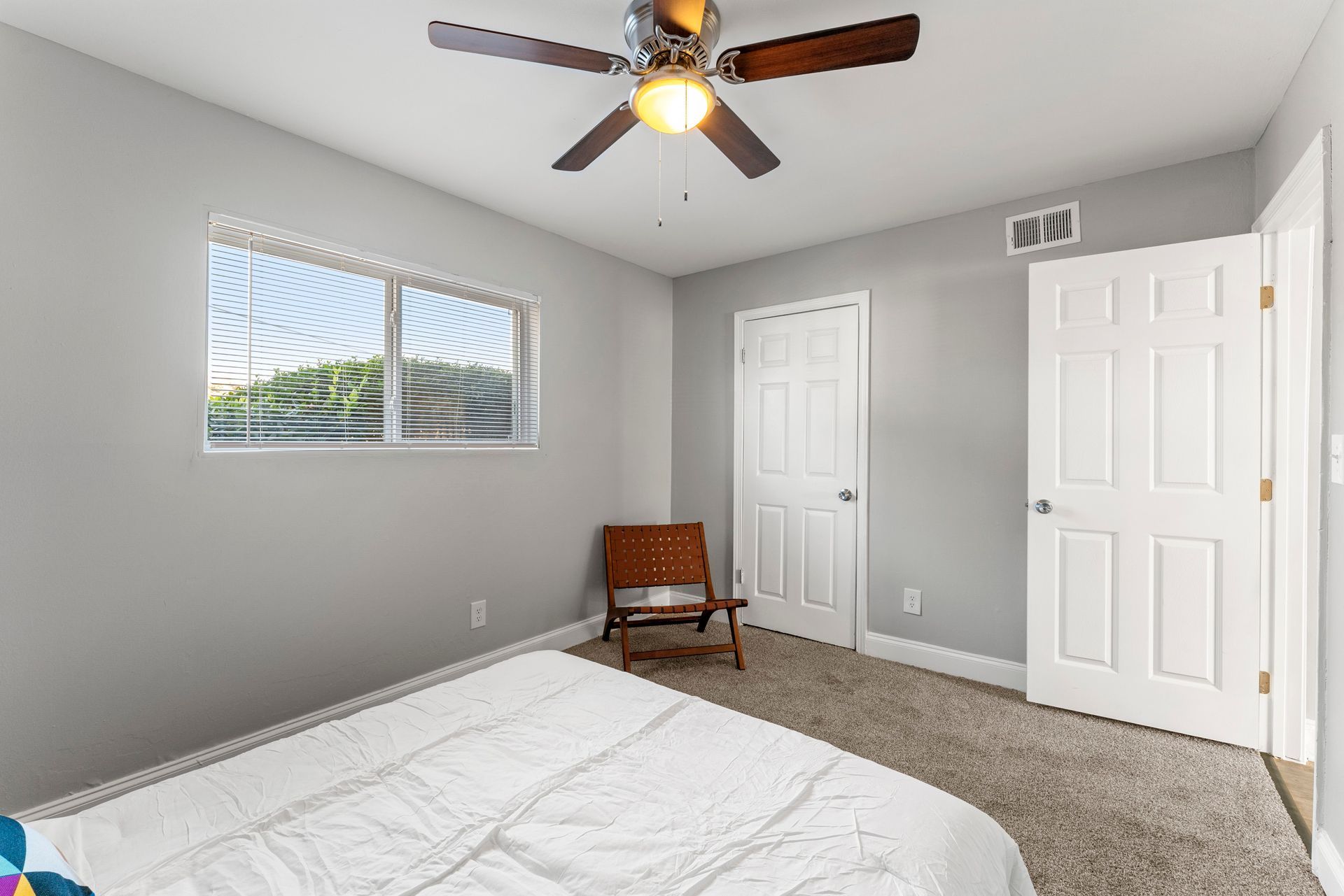Small gray bedroom with bed, ceiling fan, two white doors, chair, and window with blinds