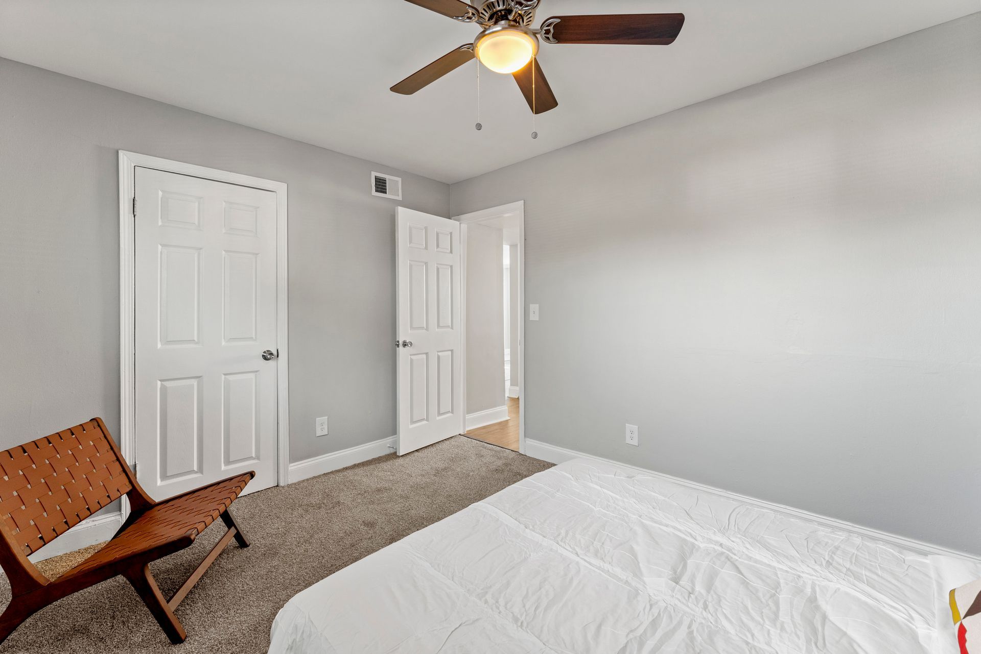 Bright bedroom with white walls, gray carpet, ceiling fan, bed, chair, and two closets with open door.