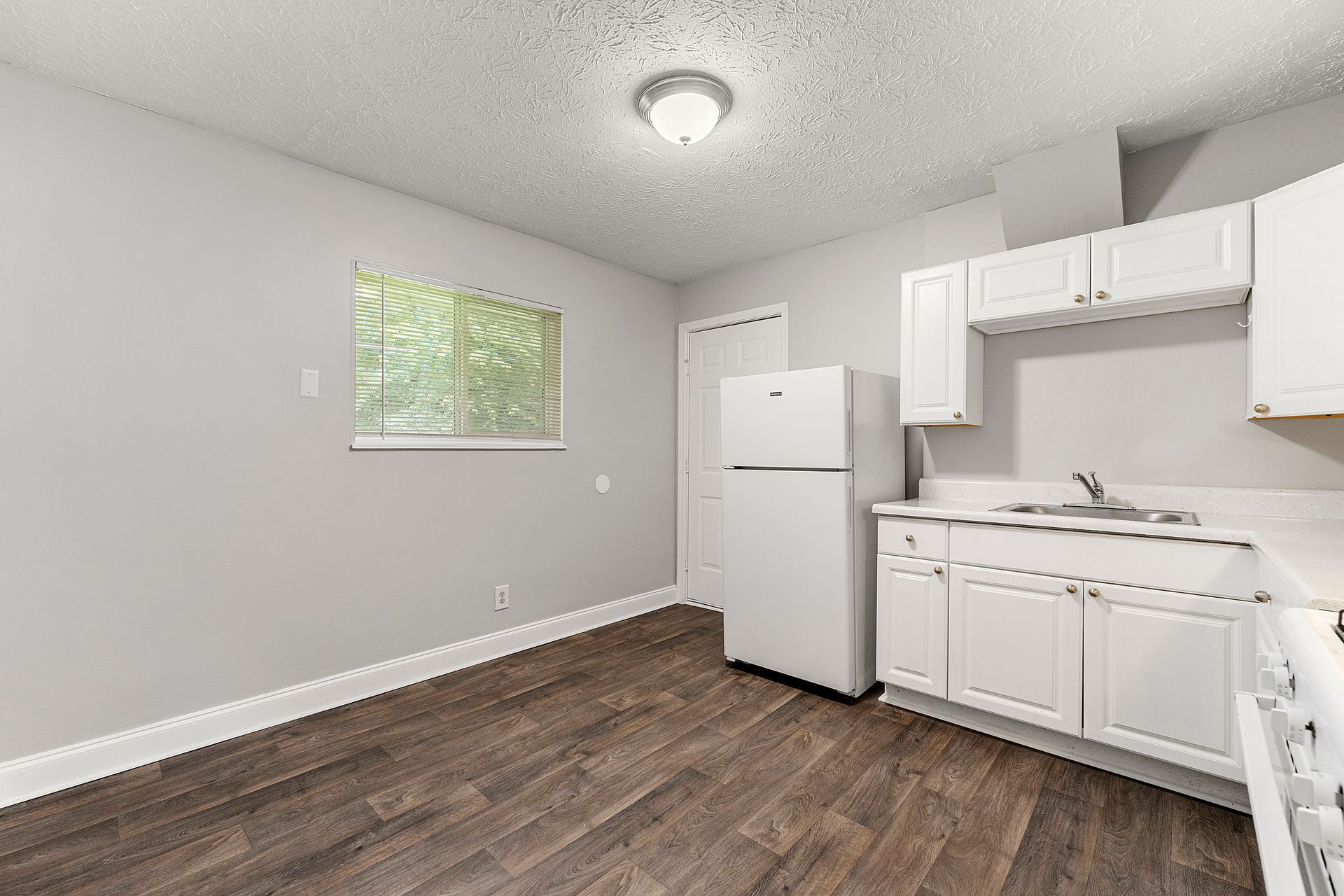 Empty kitchen with white cabinets, stove, refrigerator, and wood-look flooring in a bright room.
