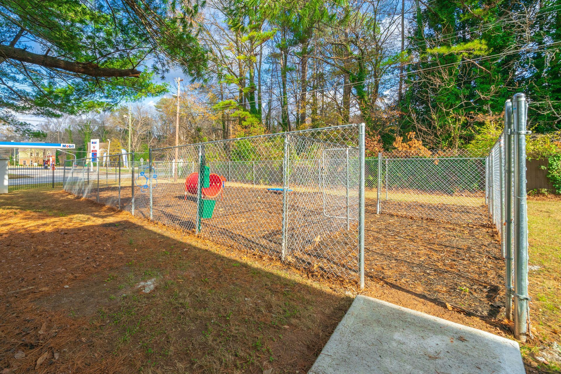 Chain-link fence around a wooded playground, with a child in red near the gate and a concrete path nearby