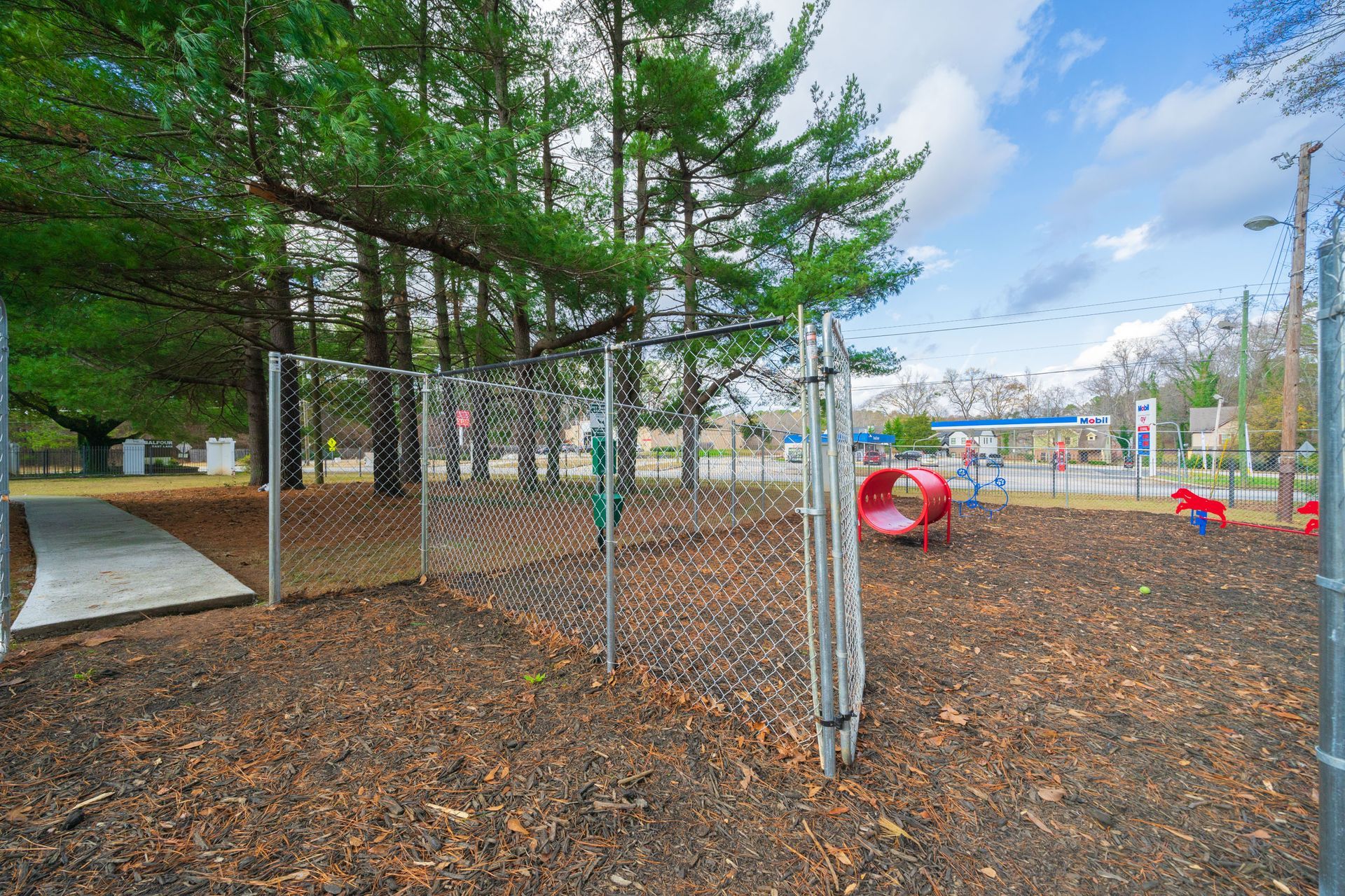 Chain-link fenced playground with red climbing tunnel, mulch ground, trees, and cloudy sky