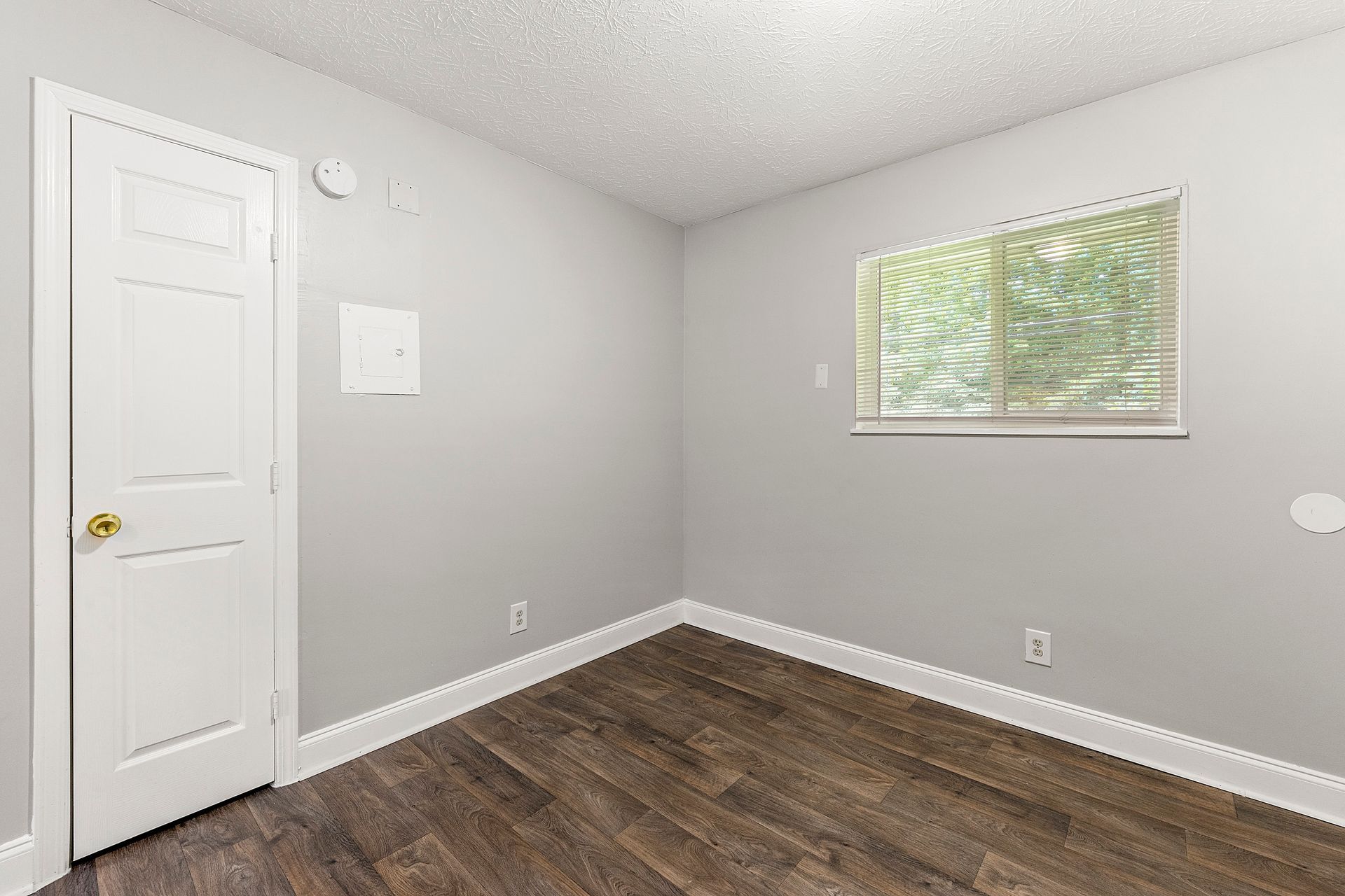 Empty gray bedroom with dark wood floor, white door, and a small window with blinds.