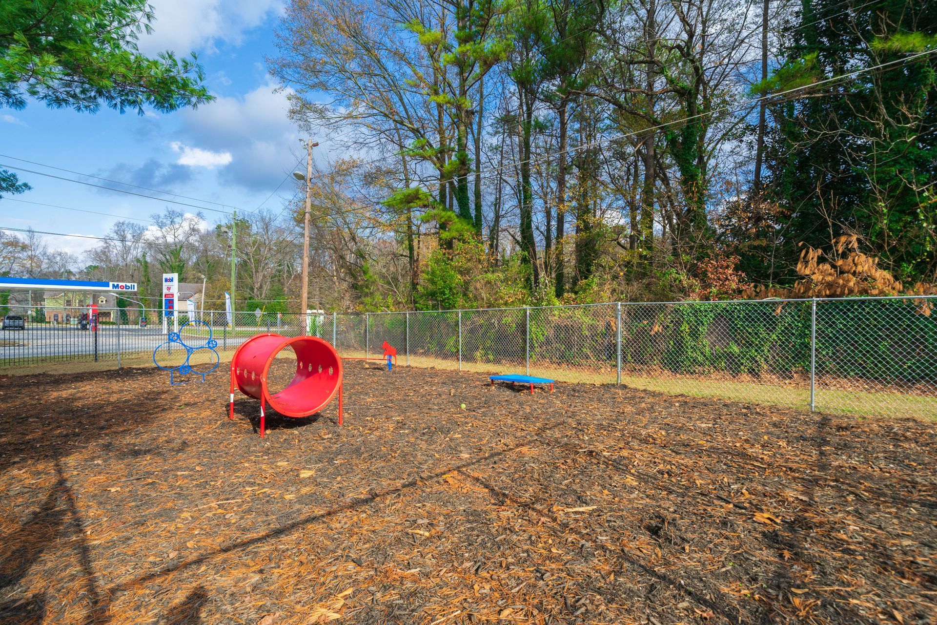 Empty playground with a red tunnel tunnel, mulch ground, chain-link fence, and trees under a blue sky