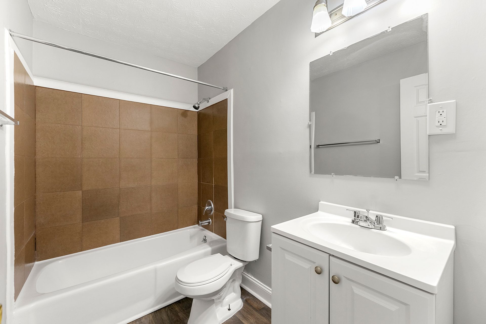 Small bathroom with tub, brown tile shower surround, white vanity, mirror, and toilet.
