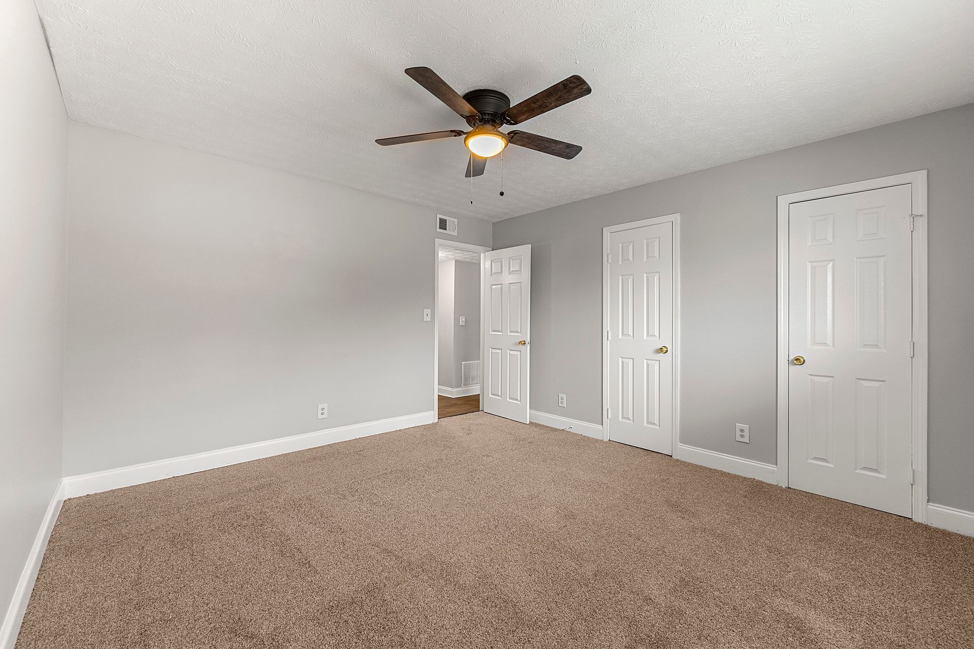 Empty beige-carpeted bedroom with gray walls, white doors, and a ceiling fan with light.