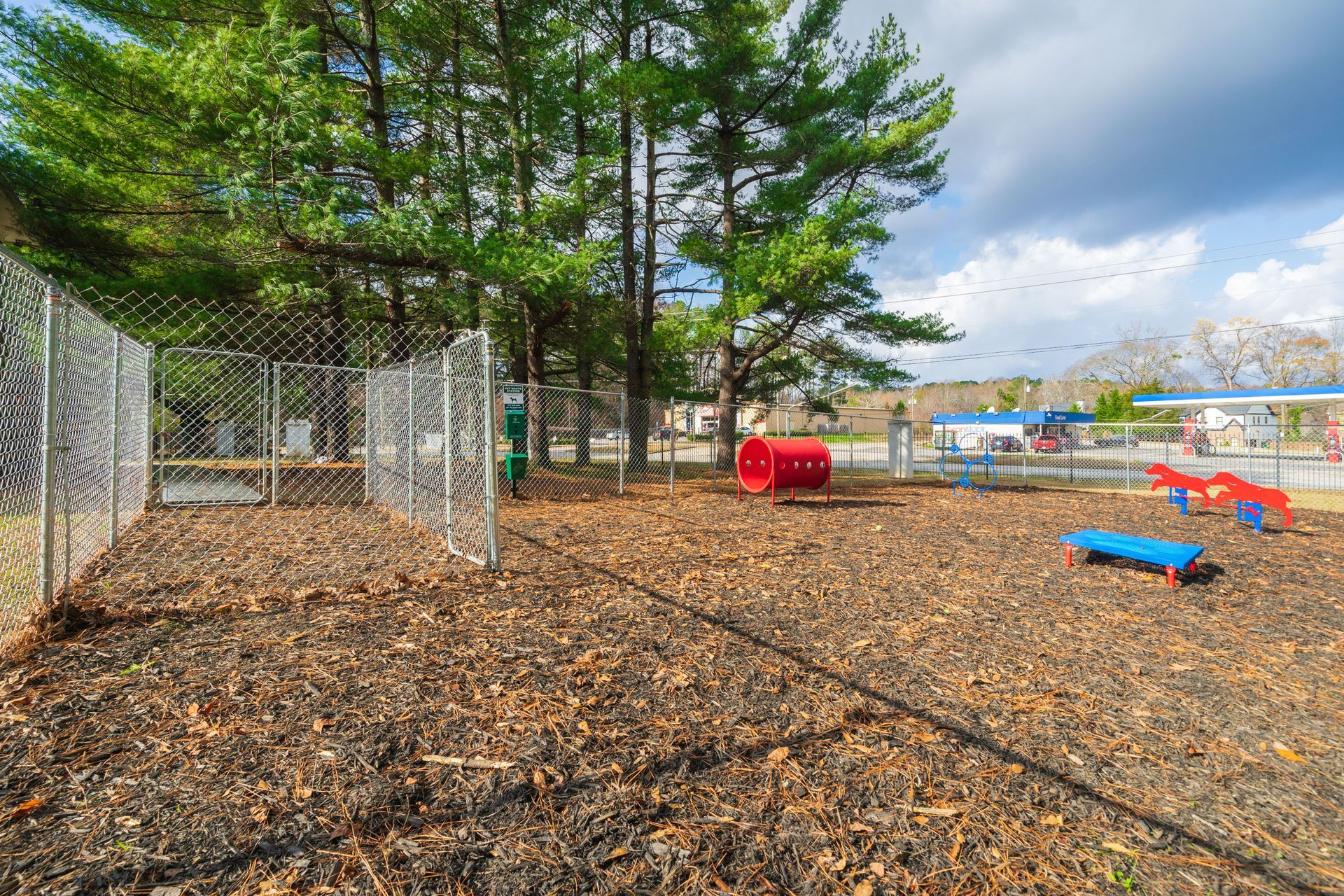Empty playground with wood-chip ground, chain-link fence, trees, and colorful play equipment under cloudy sky
