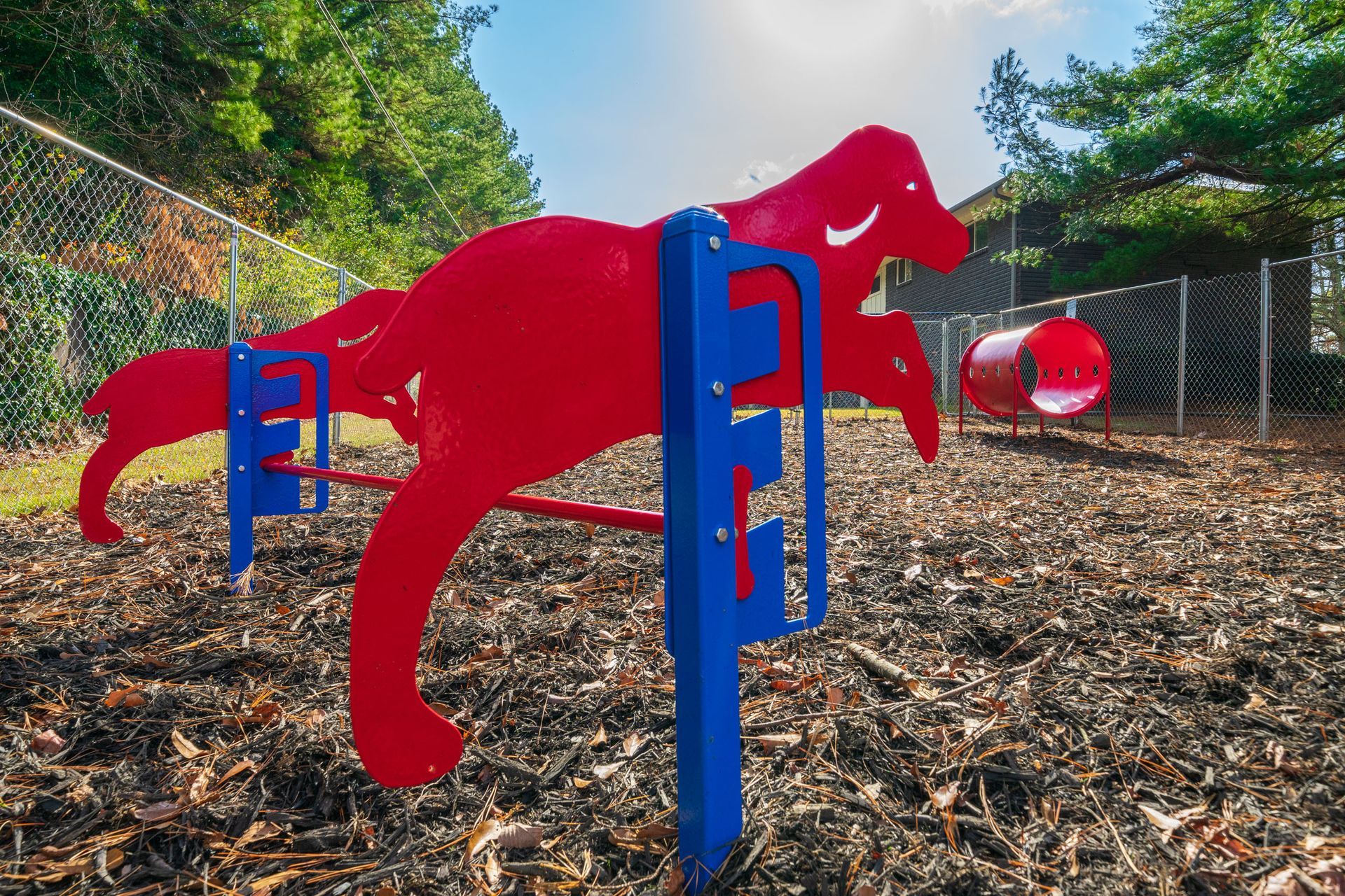 Red dog-shaped playground climbing sculpture with blue supports in a fenced outdoor area, with trees and mulch.
