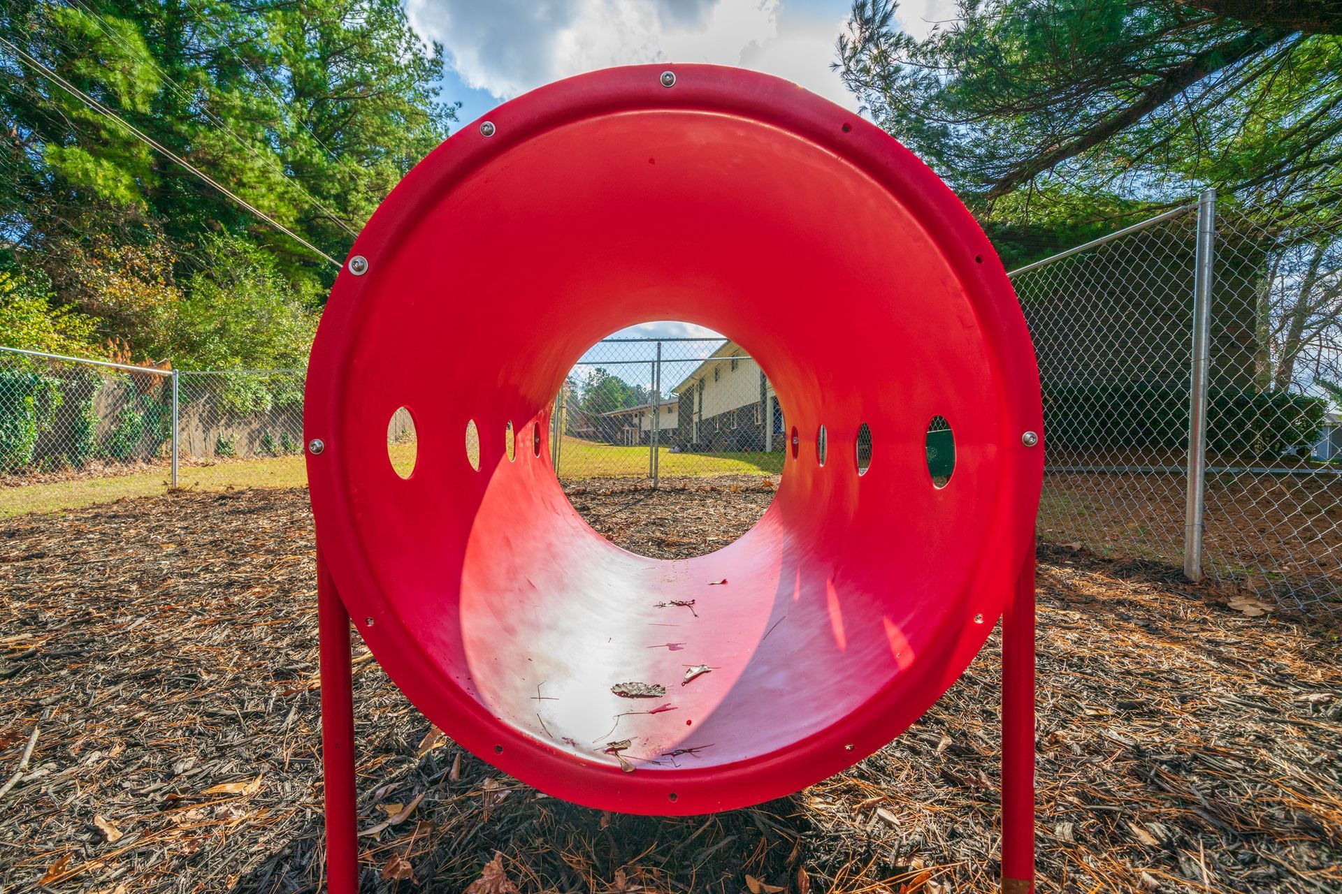 Red tunnel playground climber with round openings, set on mulch near a chain-link fence and trees.