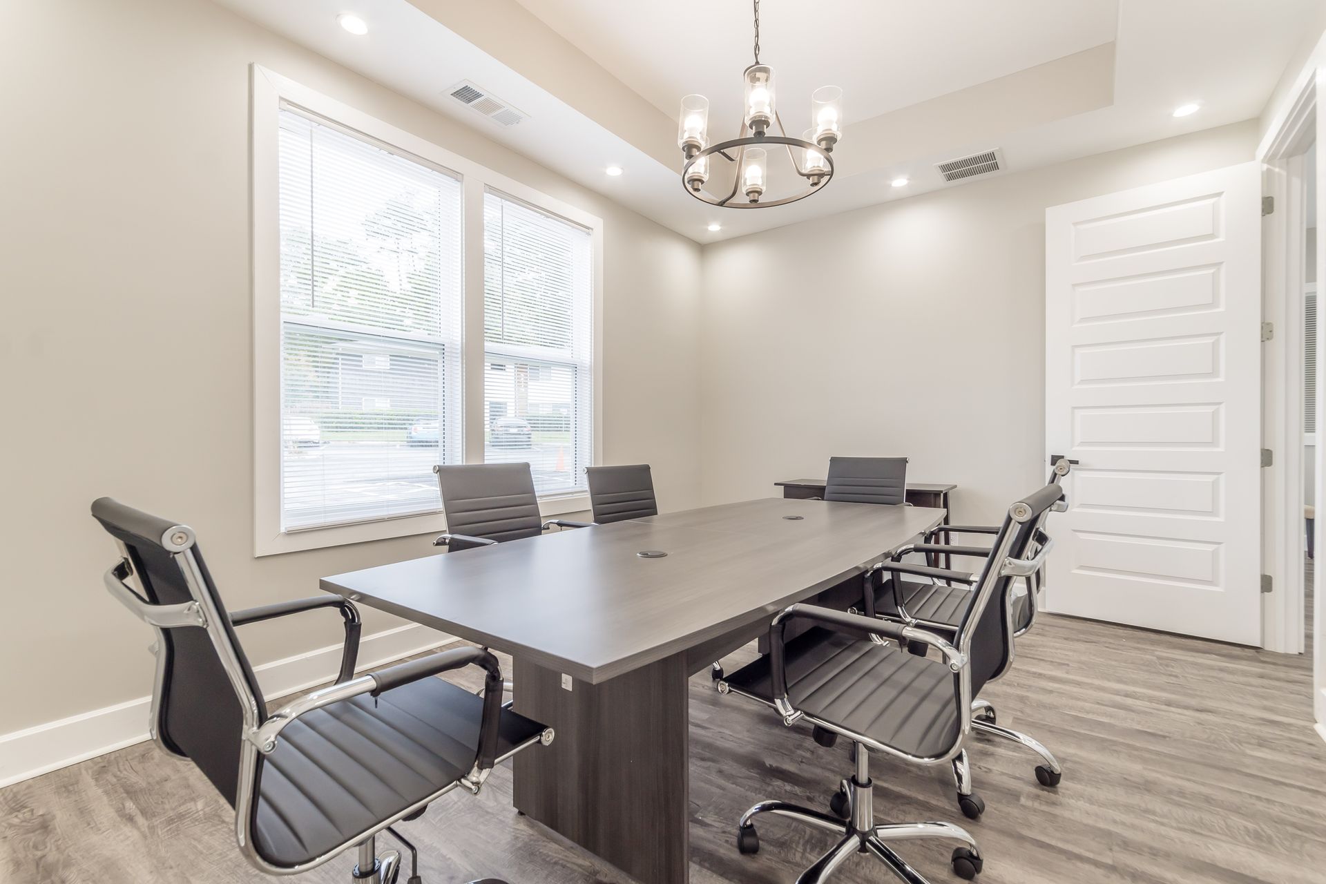 Modern conference room with long gray table, black chairs, large windows, and white door