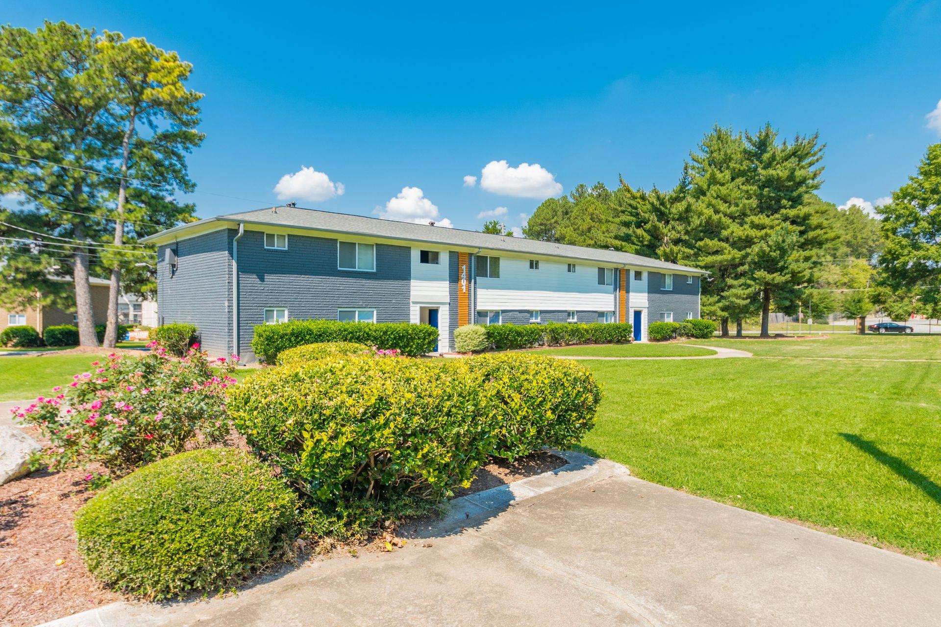 Blue apartment building with white trim, green lawn, shrubs, and a sunny sidewalk under a clear sky