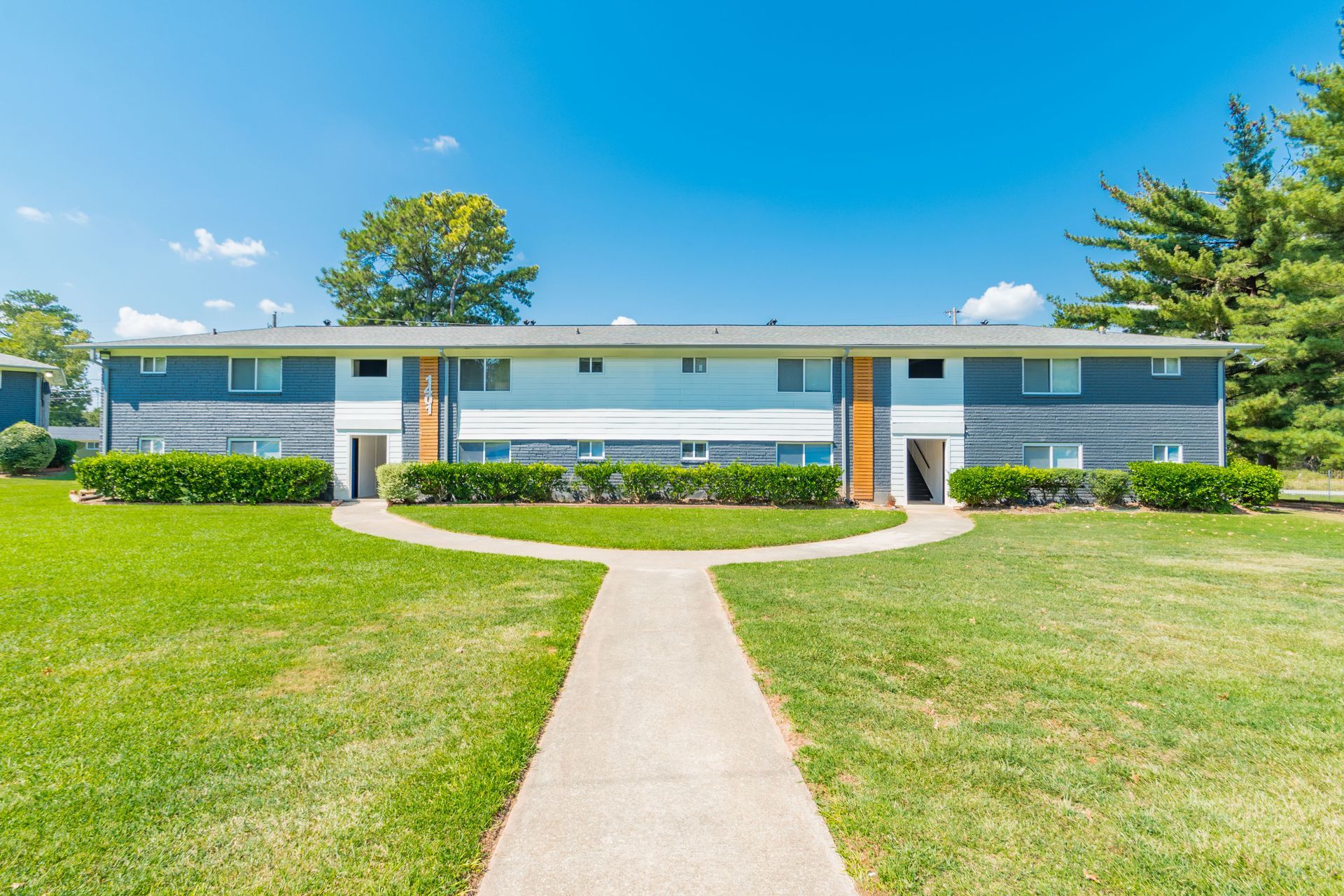 Single-story apartment building with white siding, blue trim, and a sidewalk leading through a green lawn.