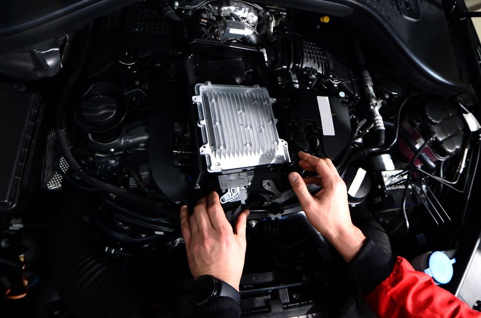 Mani che lavorano sul motore di un'auto; componenti del motore neri, rettangolo argentato, maniche della giacca rosse.