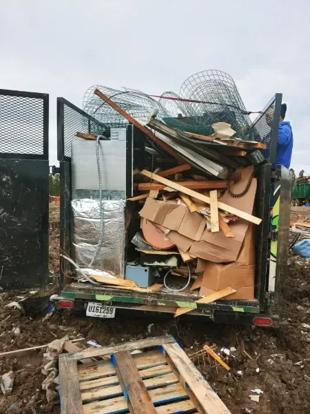 Dumpster overflowing with various debris, including wood, cardboard, and metal mesh, outdoors on muddy ground.