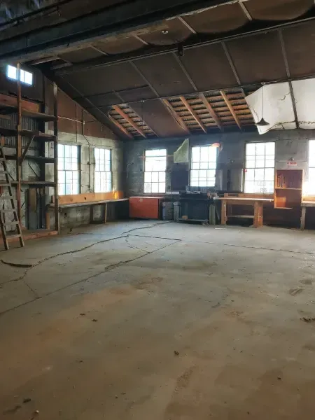 Empty workshop interior with concrete floor, workbench, windows, and exposed ceiling.
