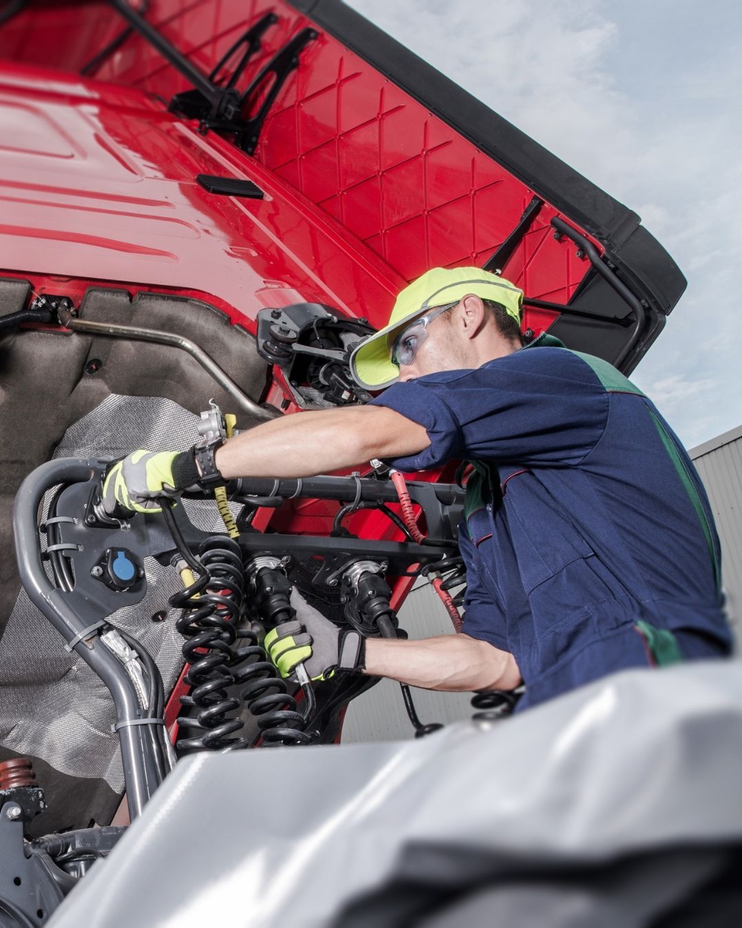 Mechanic in yellow hard hat inspects a red vehicle engine compartment under the hood.