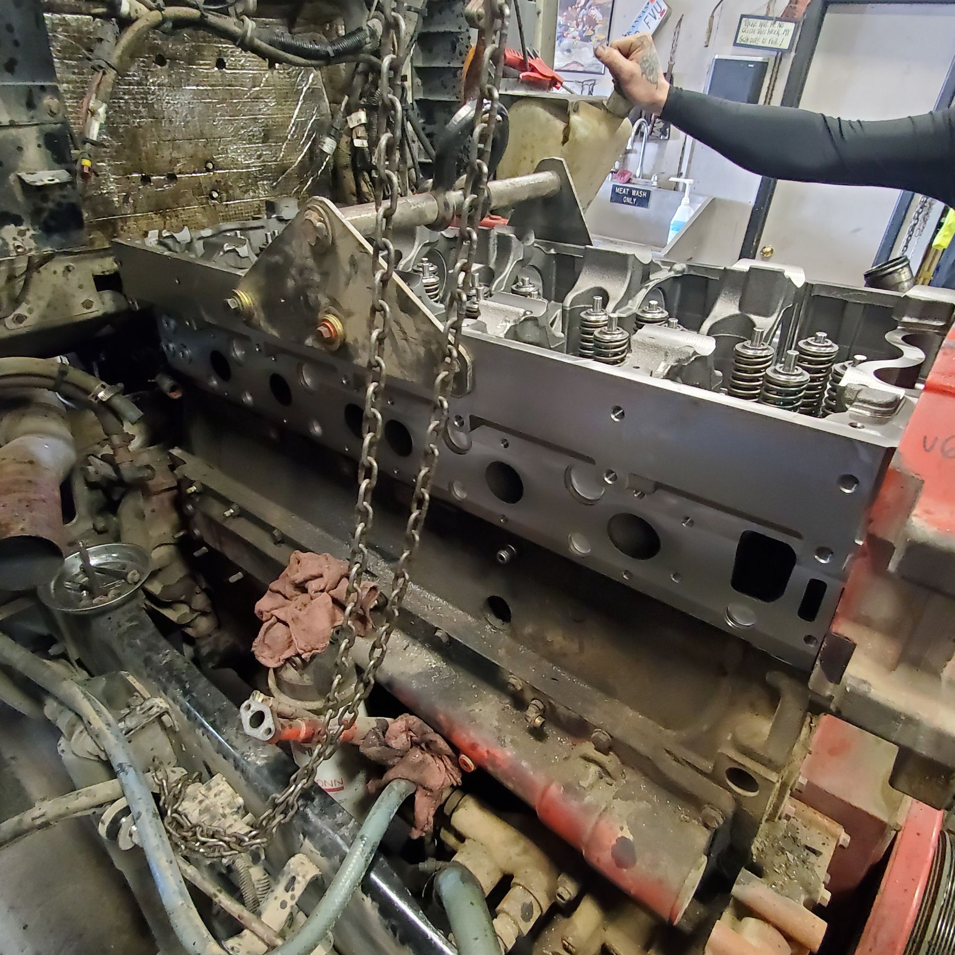 Large engine block in a garage, with chains, hoses, and a person working on the side.