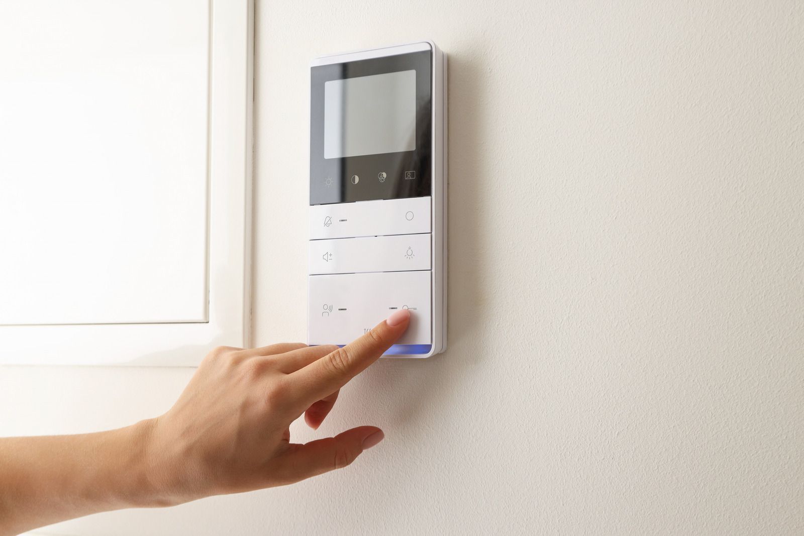 Hand pressing button on white intercom system mounted on white wall.