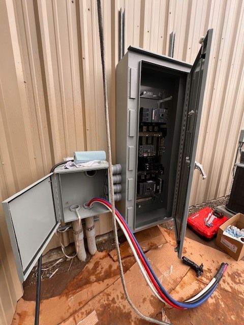 Electrical cabinet with open doors and exposed wiring on a corrugated metal building exterior.