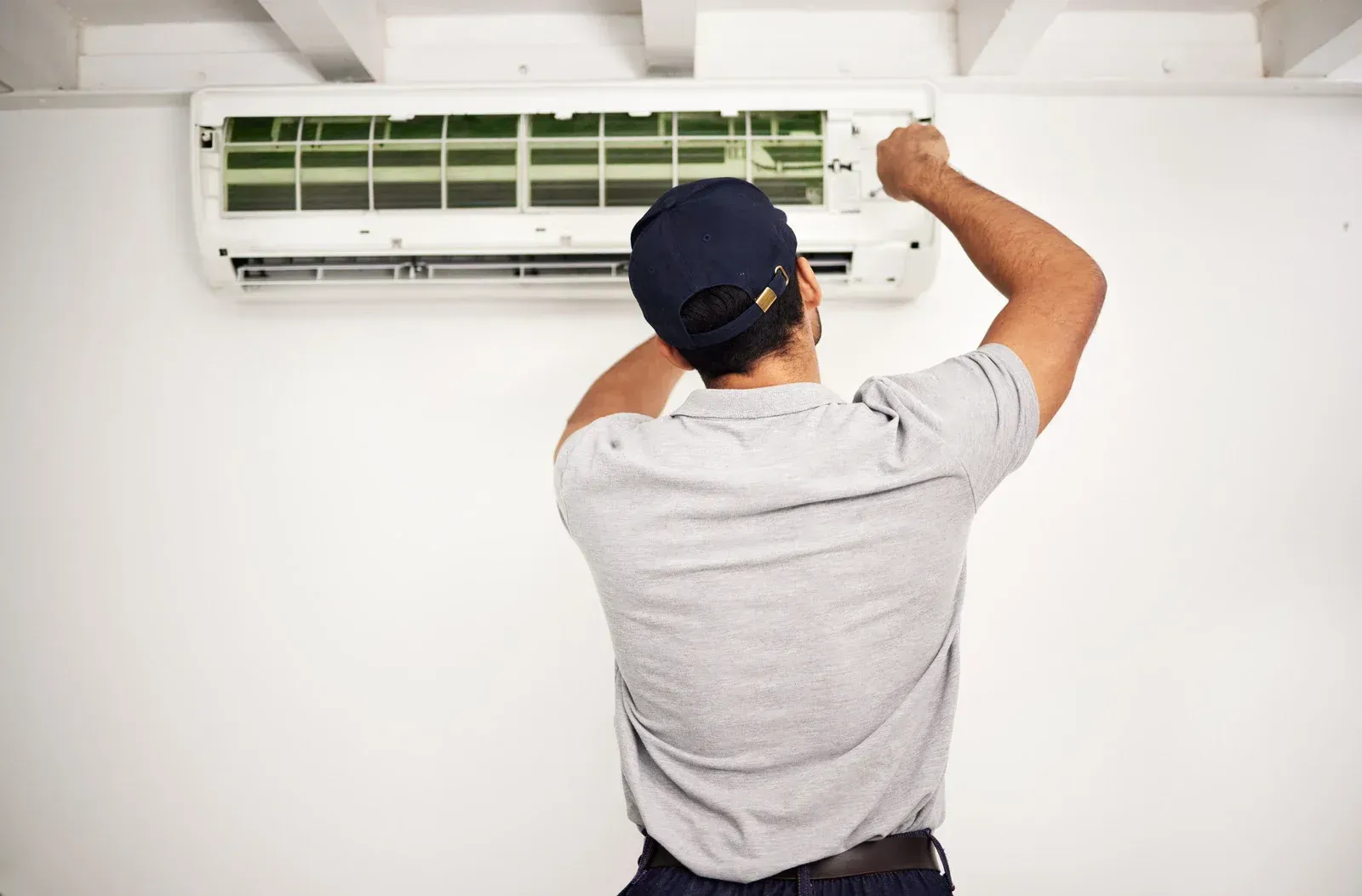 A person in a baseball cap works on a wall-mounted air conditioner in a white room.
