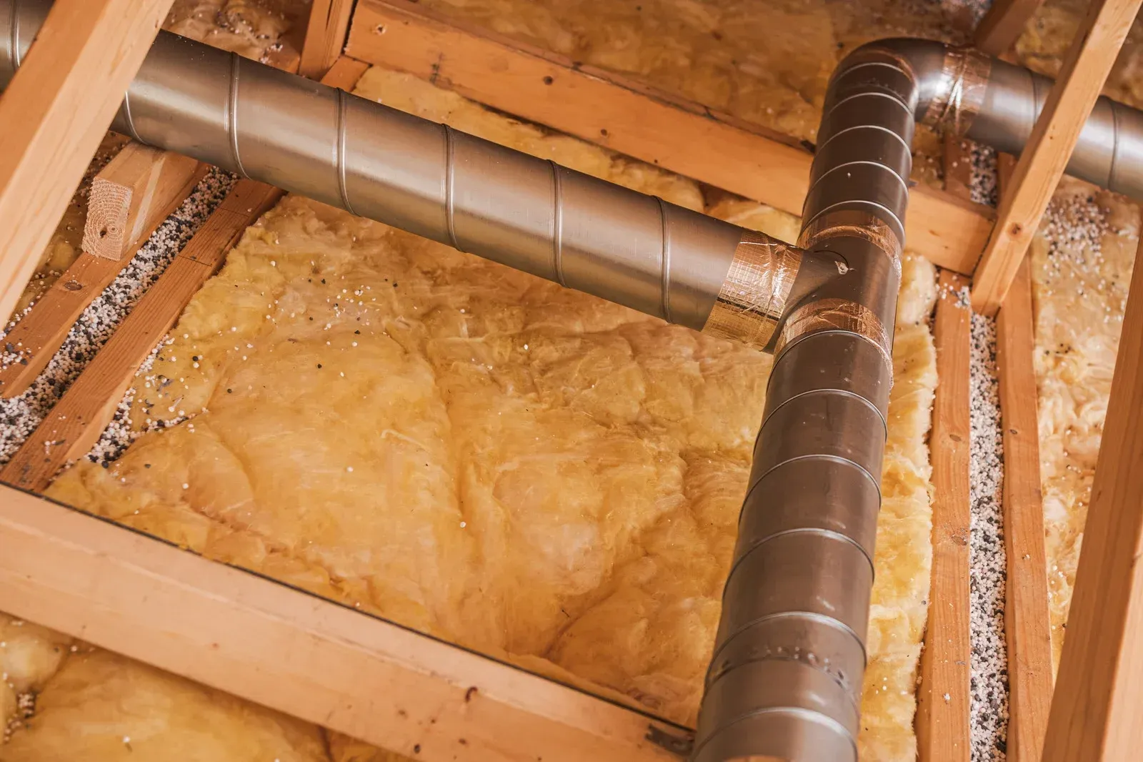 Insulation and ductwork in an attic, with wood framing and a silver metal ventilation pipe.