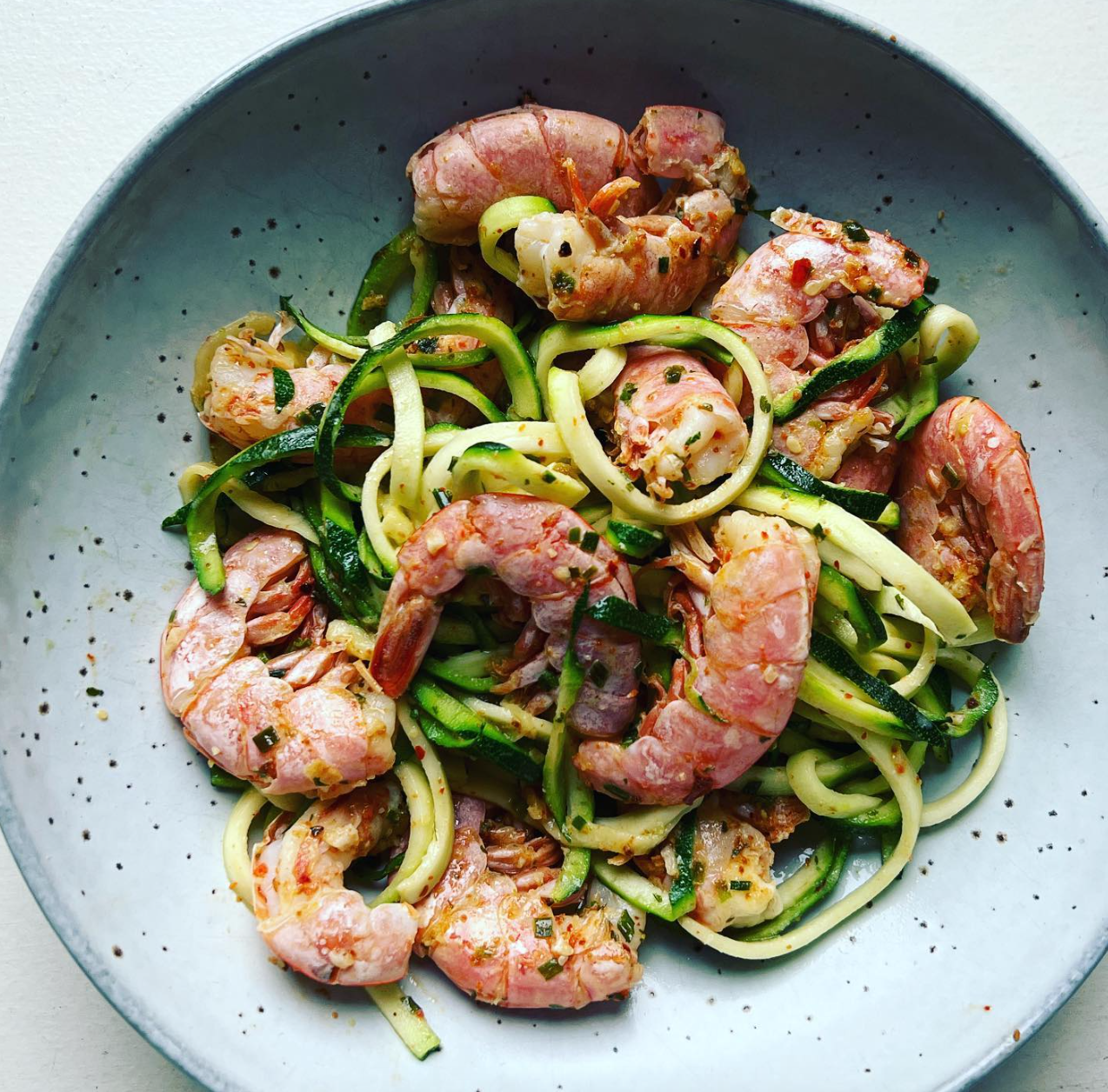 Een kom garnalen gemengd met spiraalvormige courgettenoedels, gekruid met kruiden en peper.