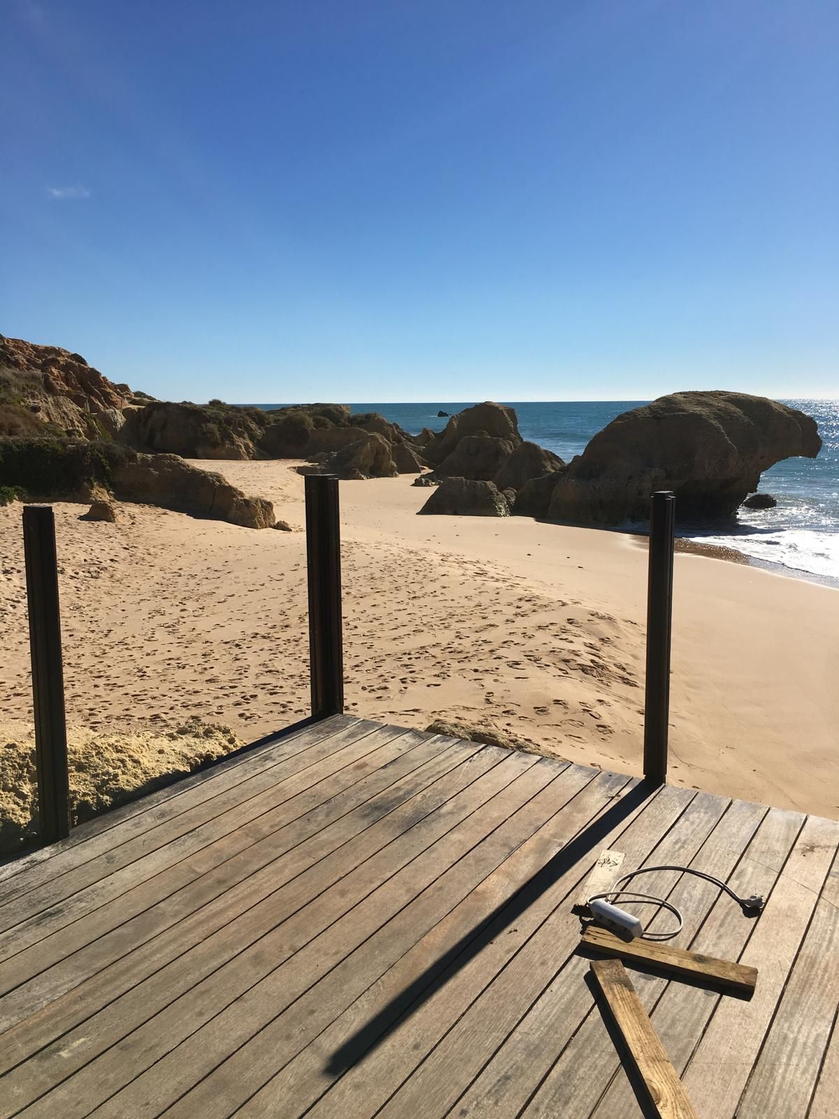 Een houten terras biedt uitzicht op een zandstrand met grote rotsformaties onder een helderblauwe hemel.