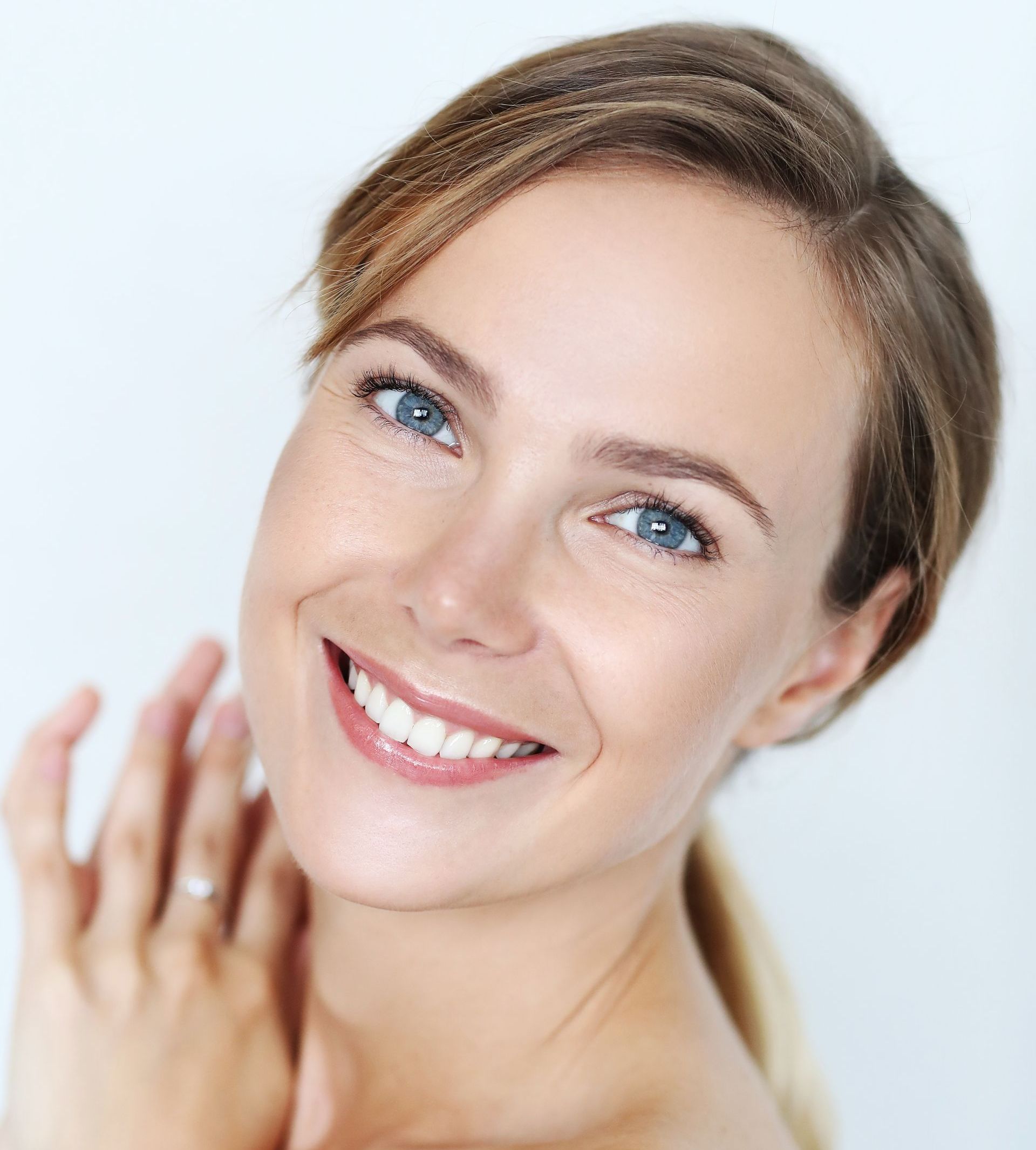 A woman with blue eyes is smiling and touching her face.