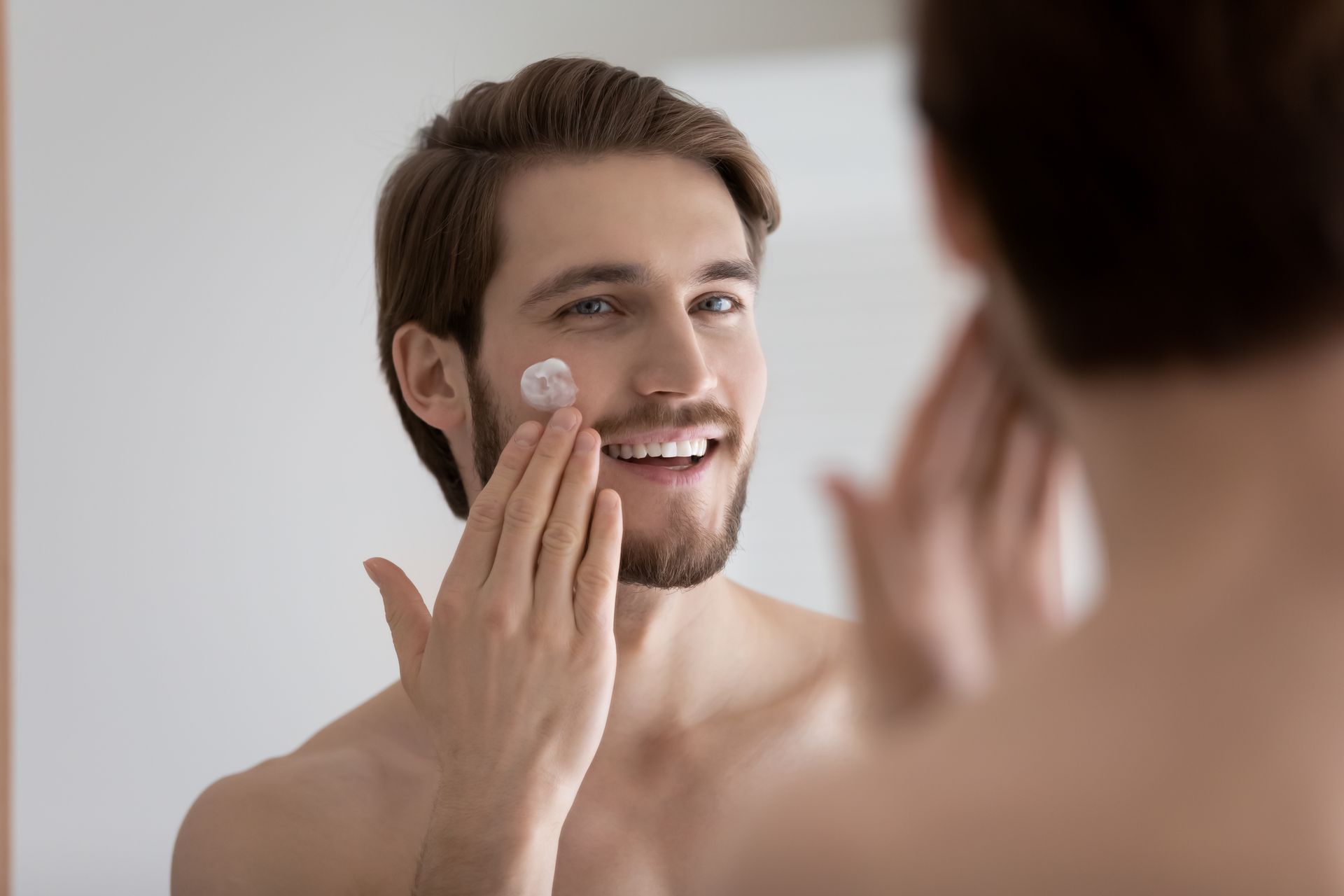 A man is applying cream to his face in front of a mirror.