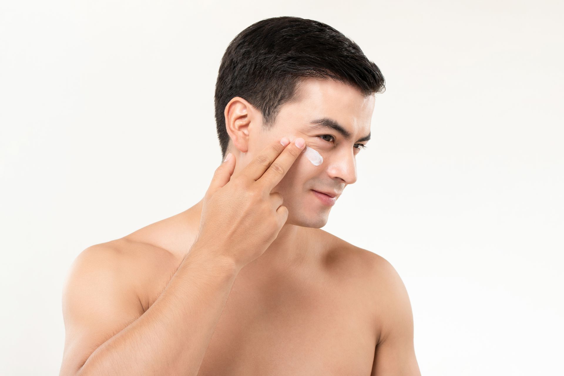 A shirtless man is applying lotion to his face.