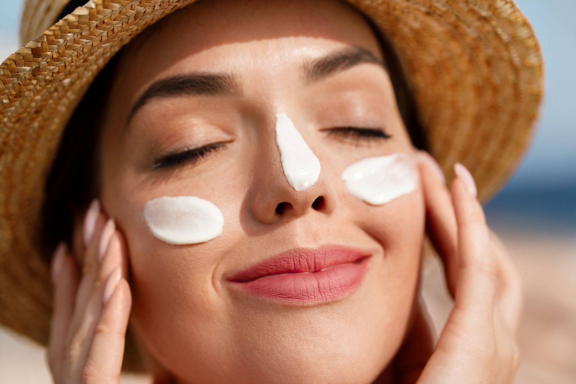 A woman wearing a hat is applying sunscreen to her face.