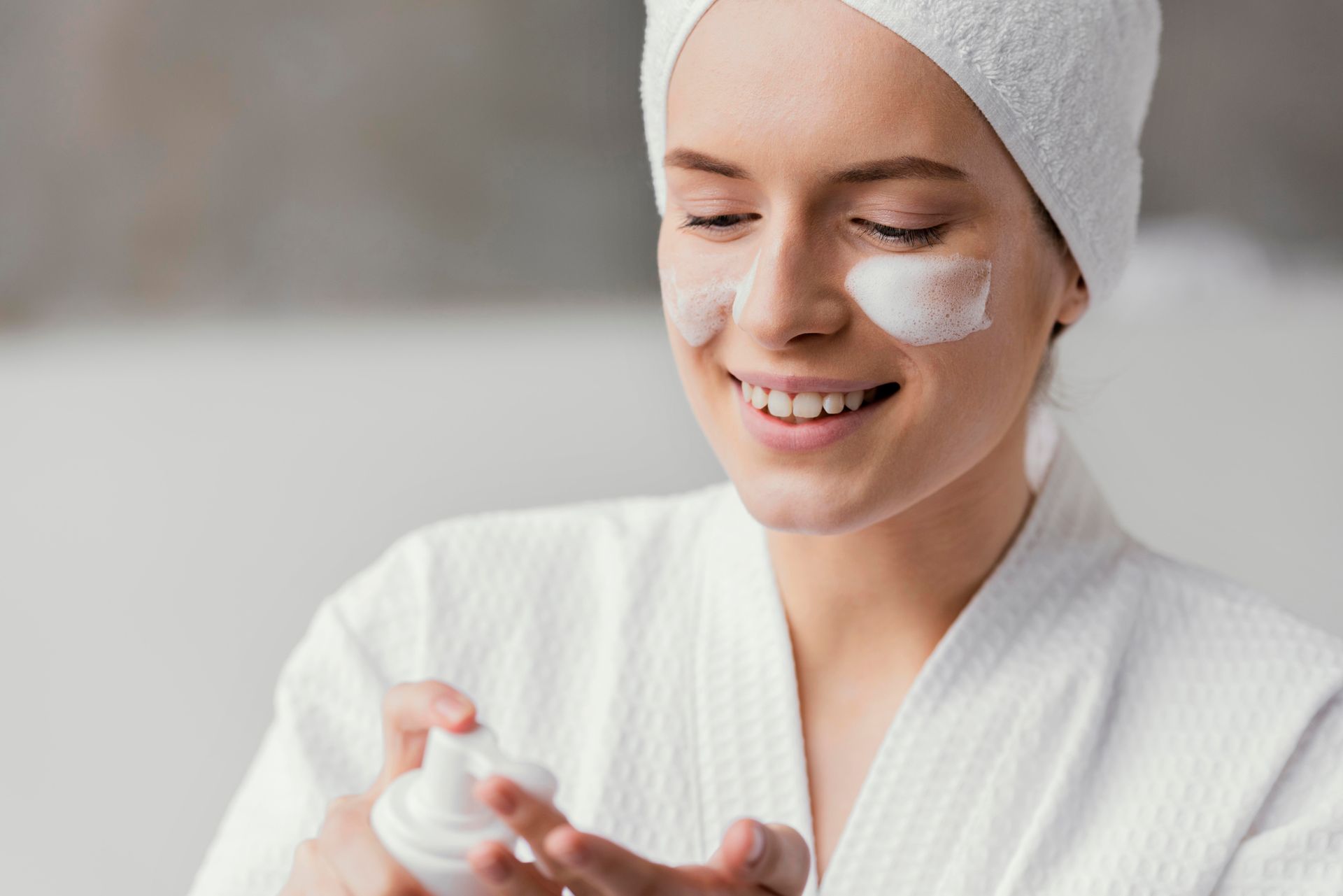 A woman with a towel wrapped around her head is applying lotion to her face.