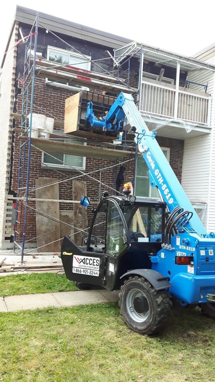 Un chariot télescopique bleu soulève du bois près d'un bâtiment en briques en cours de rénovation. On aperçoit un échafaudage et de l'herbe.