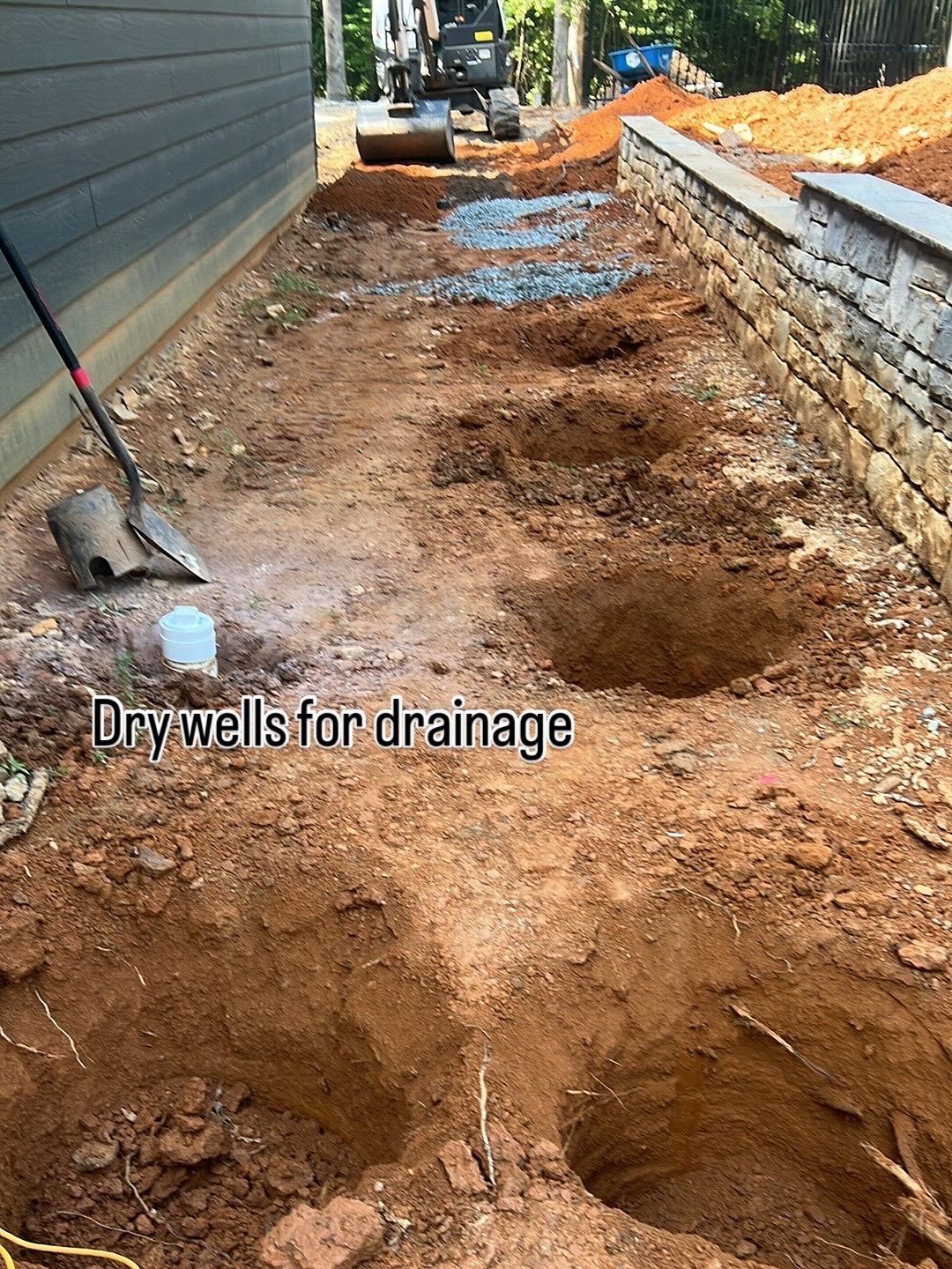 Construction site with excavated holes for dry well drainage; a building and retaining wall are visible.