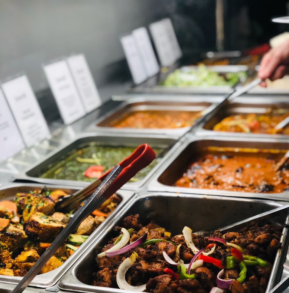 Buffet with various Indian dishes in serving trays; person using tongs.