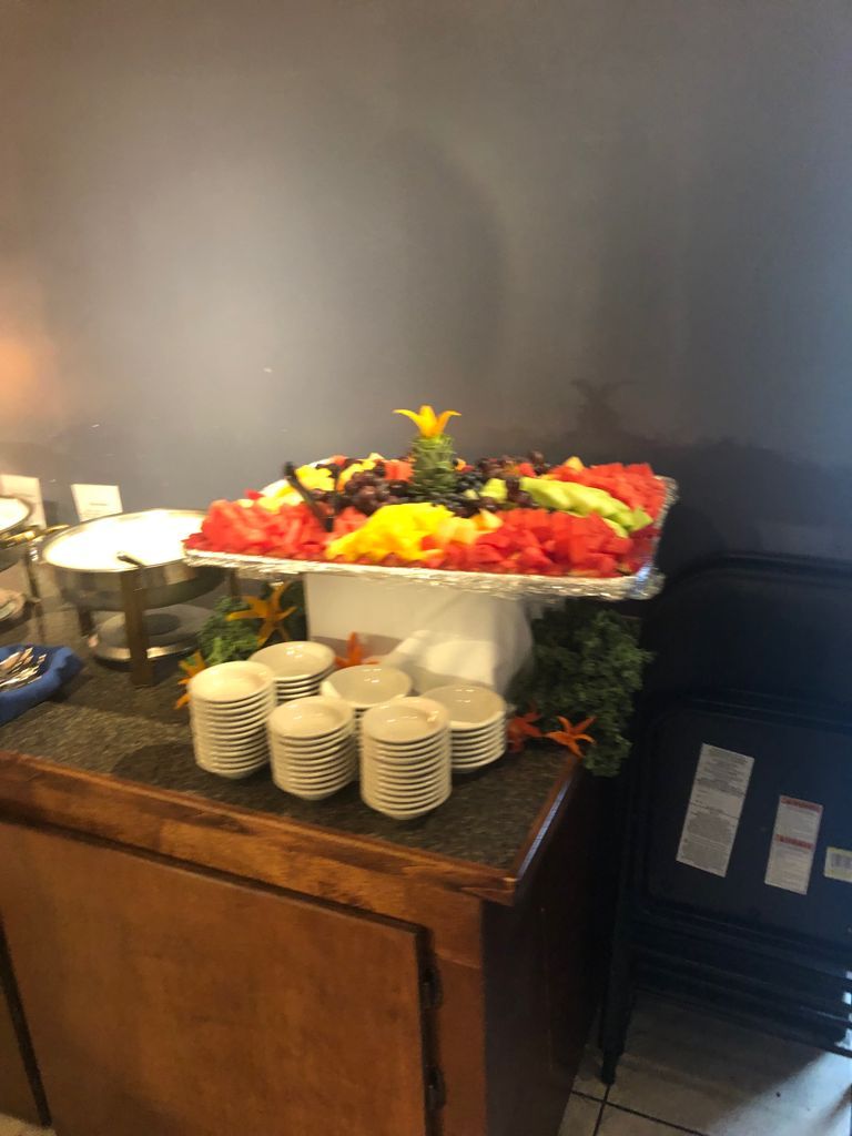 Fruit platter on a table with cups, next to a stainless steel chafing dish and a stack of black chairs.