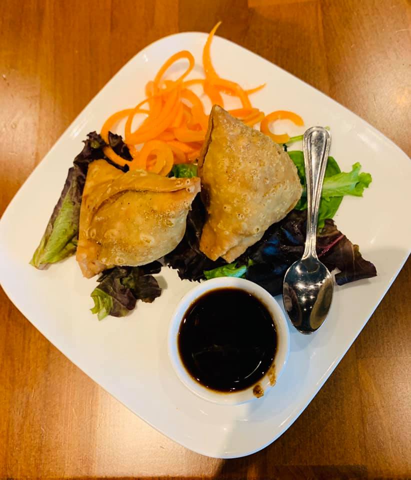 Two golden samosas on a plate with greens, shredded carrots, and dark dipping sauce, with a spoon.