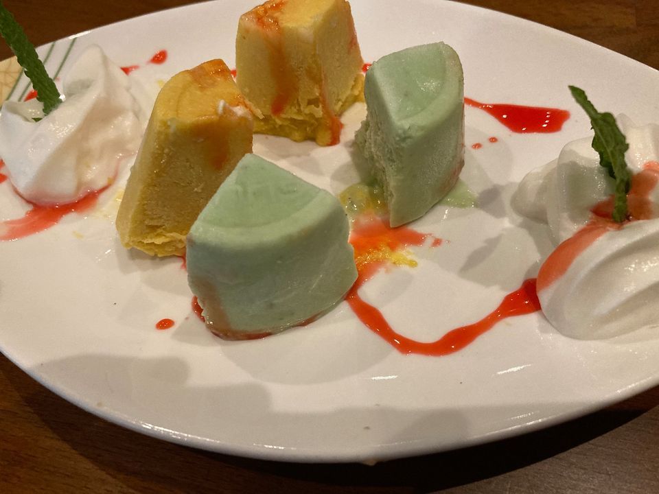 Ice cream dessert with green, yellow, and white treats, drizzled with red sauce and whipped cream on a white plate.