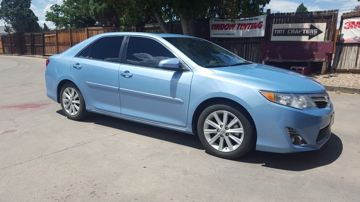 Light blue sedan parked on a sunlit street near a fence and trees