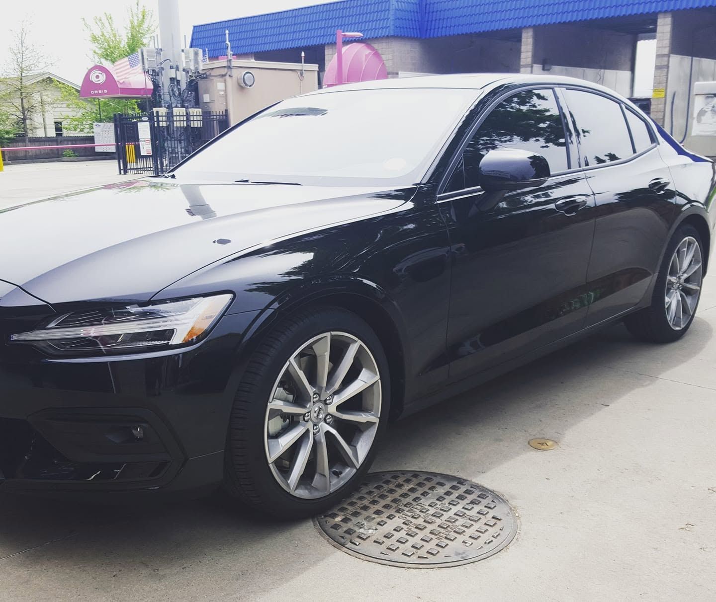 Black sedan parked on a street beside a building, with silver wheels and reflections on the hood.