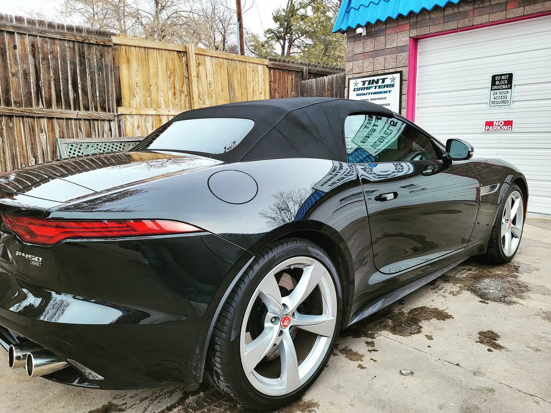 Black convertible sports car parked outdoors beside a fence and sign, viewed from the rear.