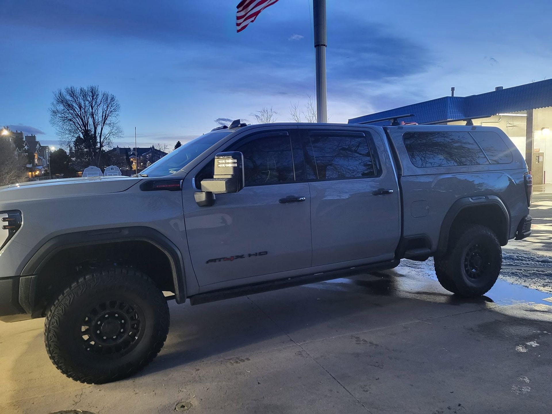 Gray pickup truck parked in a snowy lot at dusk, with an American flag behind it