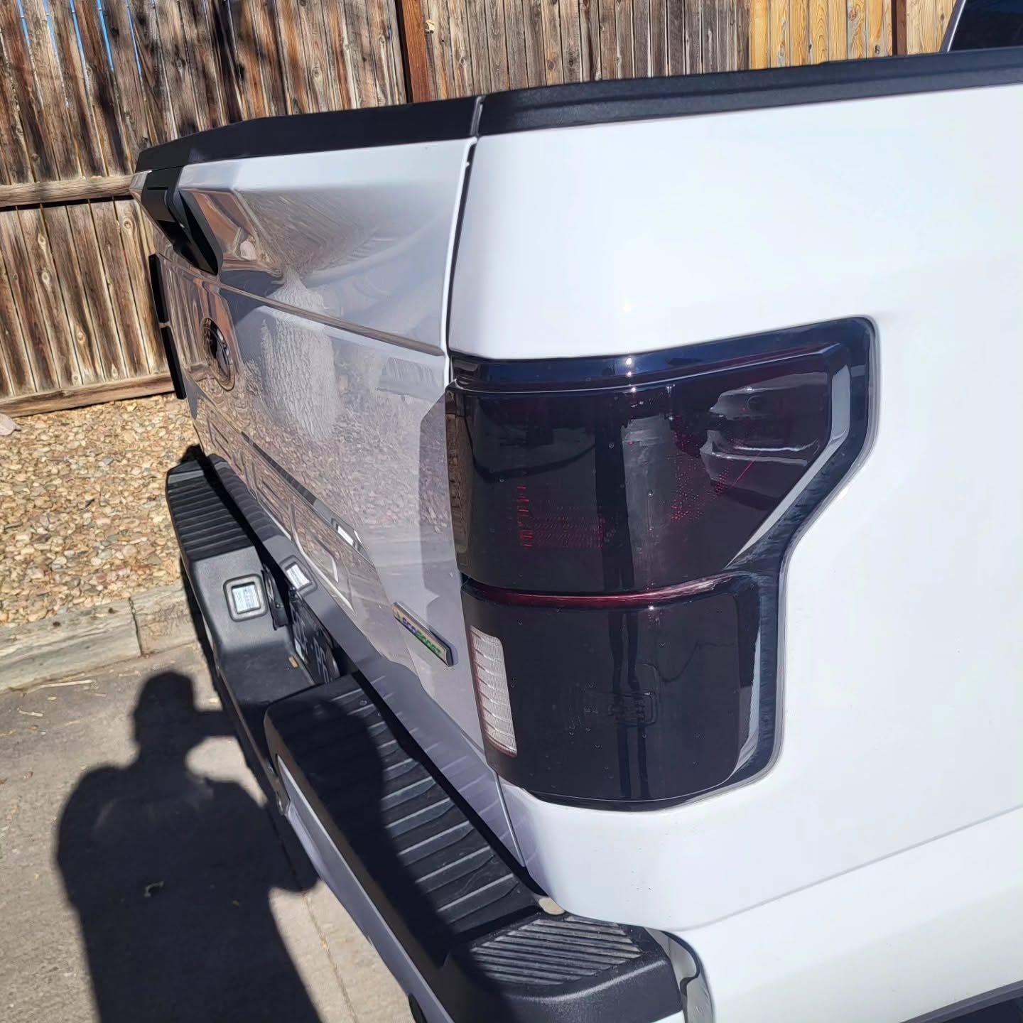 White pickup truck rear corner with tinted taillight and bed cover, parked beside a wooden fence.